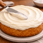 Powdered sugar frosting being smoothed over a golden cake with an offset spatula, creating soft swirls and an even, creamy finish.